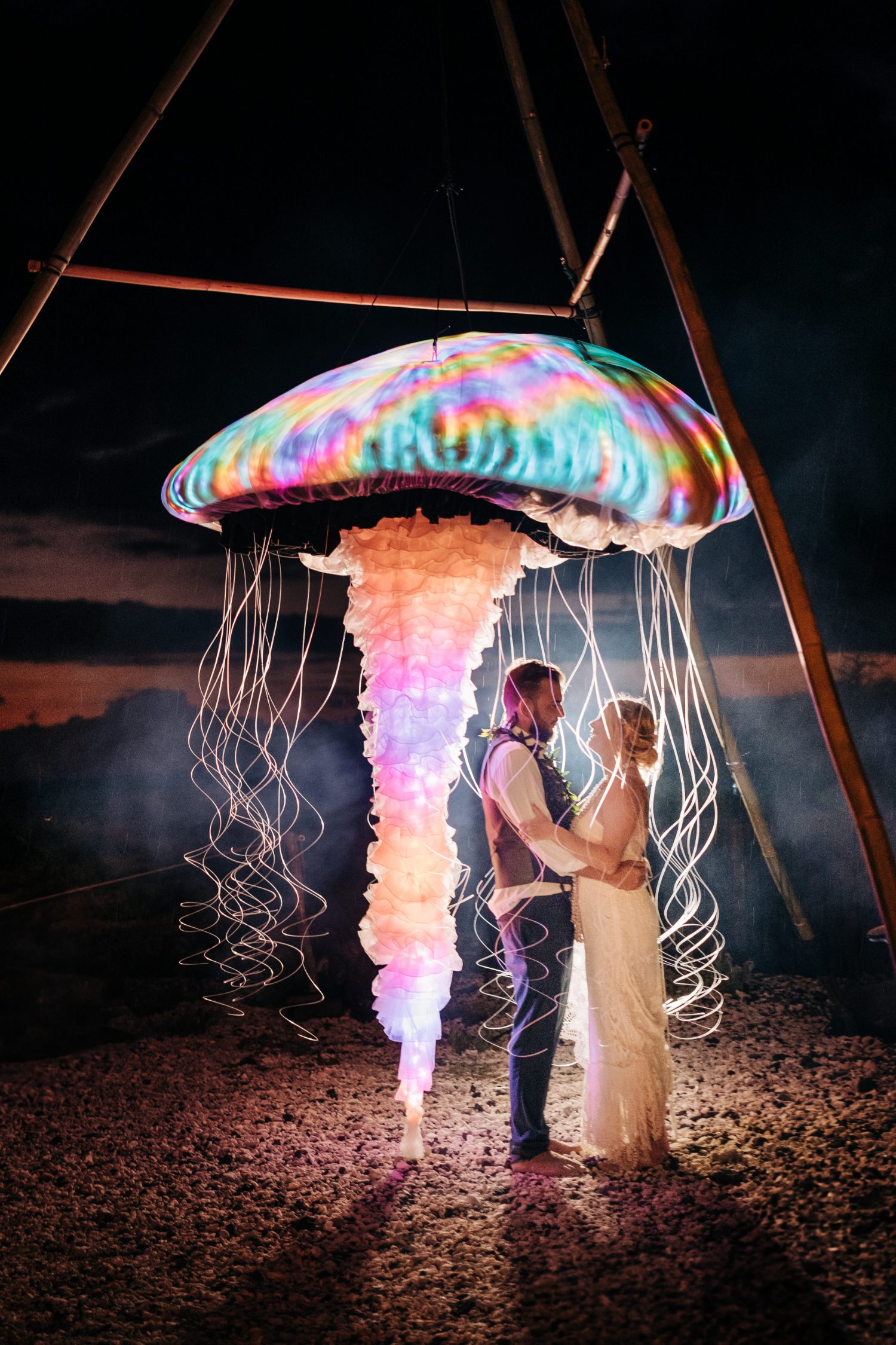 Bride and groom beneath a rainbow-lit LED jellyfish sculpture at a Kona Salt Farm wedding on the Big Island